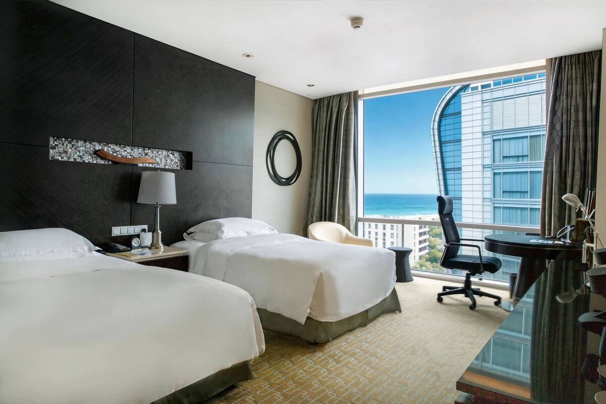 A hotel room with two beds, a desk, a chair, a lamp, and a view of the ocean.