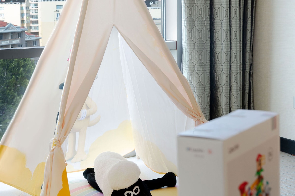 A room with a view of buildings through a window, a white teepee with a label, a black plush toy, and a colorful box on a table.