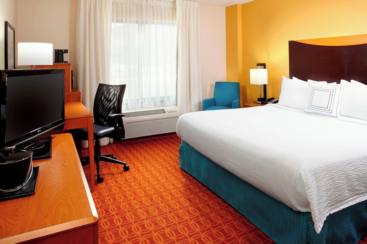 Une chambre d’hôtel avec un lit, un bureau avec une télévision, une chaise, une lampe et une fenêtre avec des rideaux.