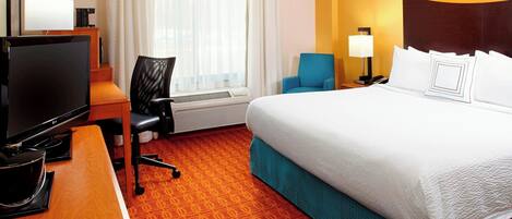 A hotel room with a bed, a desk with a TV, a chair, a lamp, and a window with curtains.