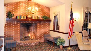 A cozy room with a brick fireplace, a chandelier, and a flag. There are two armchairs and a table with books.