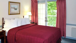 A hotel room with a large bed, a nightstand, a lamp, a framed picture on the wall, and a view of greenery through the window.