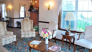 A room with a patterned carpet, floral armchairs, a wooden coffee table with a magazine, a vase with flowers, and a view of greenery through a window.