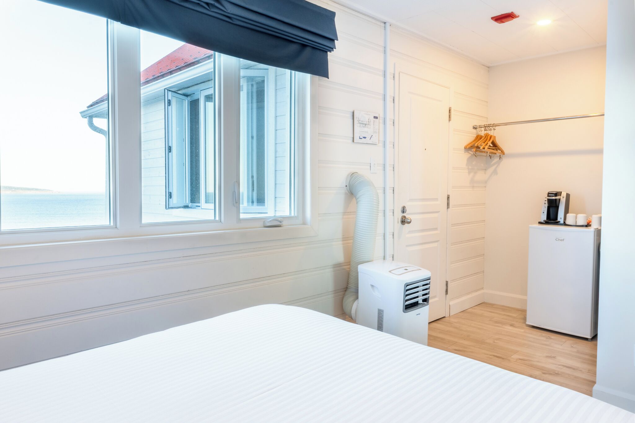 A hotel room with a bed, a window with a view, a heater, a refrigerator, and a coat rack.