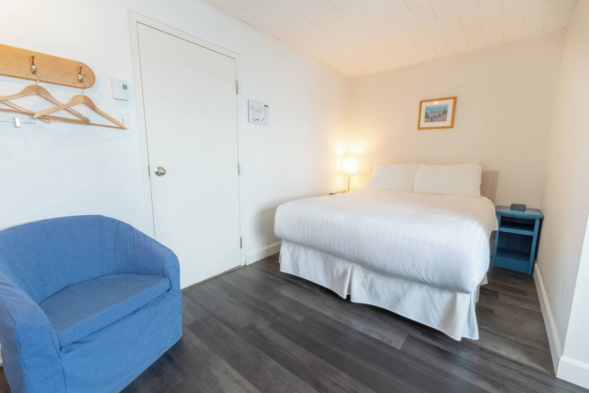 Habitación de hotel con una cama, un sillón azul, una mesita de noche y una lámpara de pared.