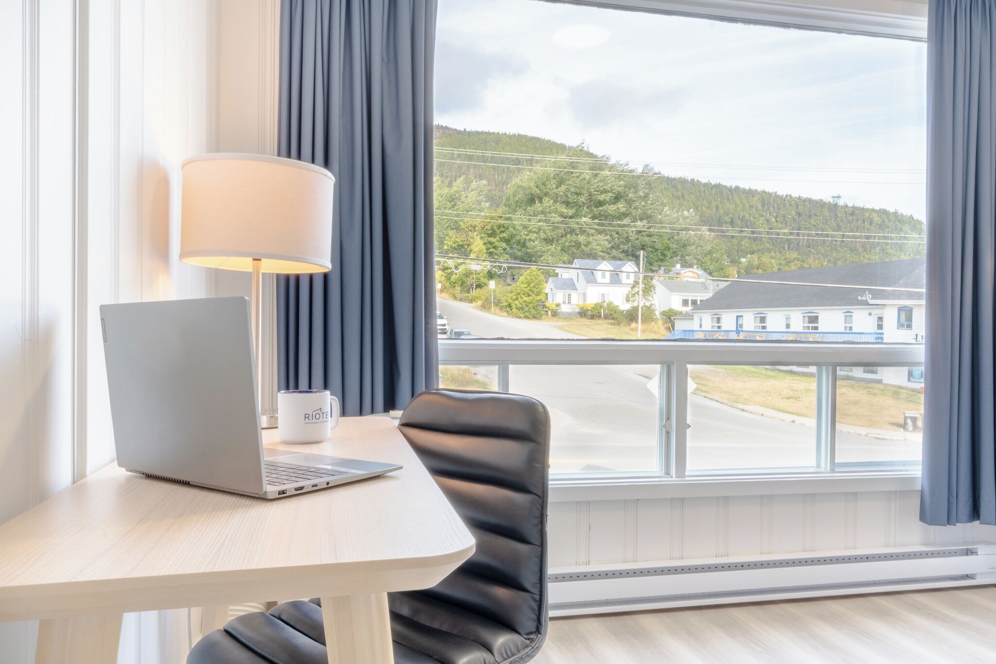 A room with a laptop on a table, a chair, a lamp, and a view of a road and buildings through a window.