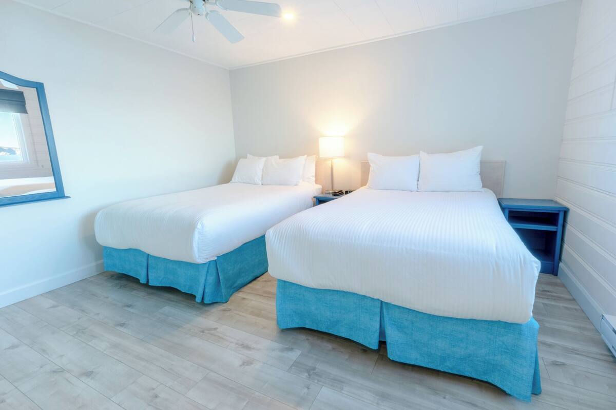 Habitación doble con dos camas, cada una con falda azul, ropa de cama blanca y mesita de noche con lámpara.