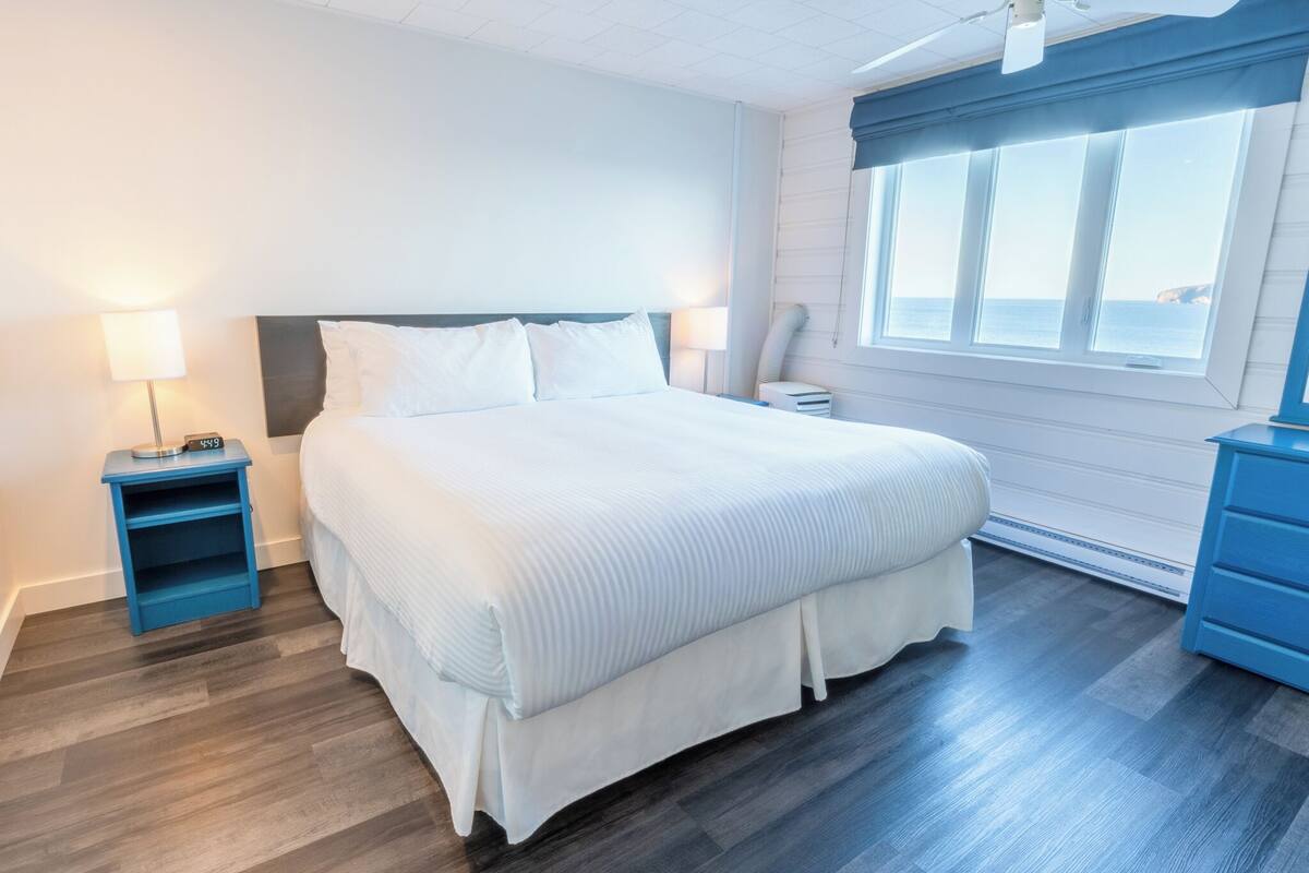 Una habitación de hotel con una cama grande, dos mesitas de noche, un tocador, un aparador y una ventana con vista al mar.