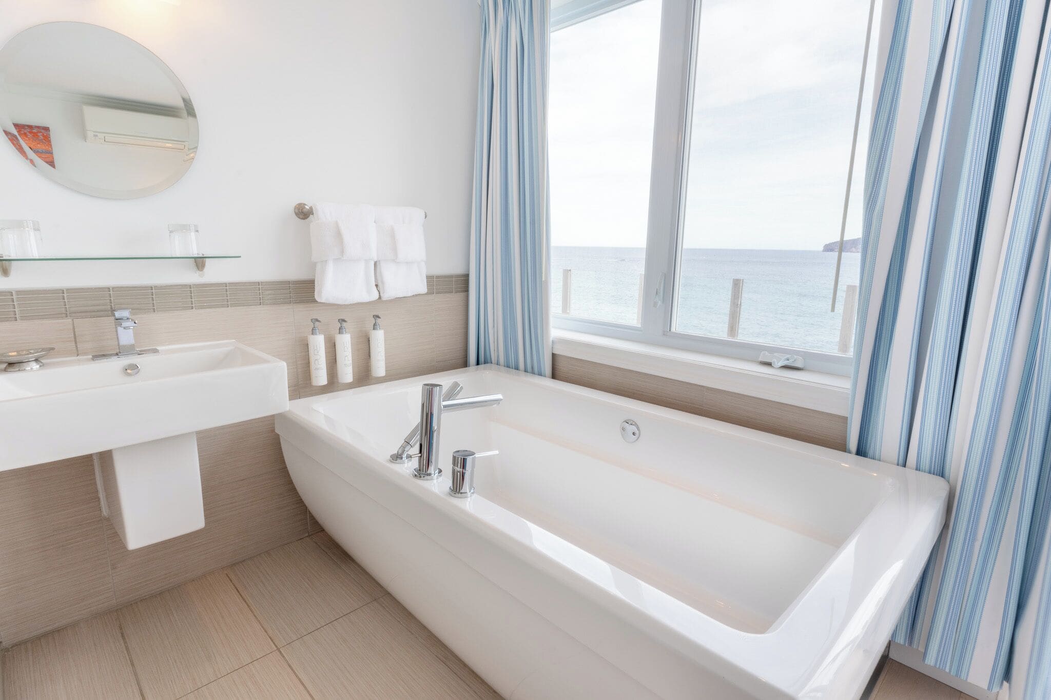 Ein modernes Badezimmer mit einer großen Badewanne, einem Waschbecken und einem Spiegel. Ein Fenster mit Vorhängen bietet Blick auf das Meer.