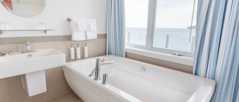 Ein modernes Badezimmer mit einer großen Badewanne, einem Waschbecken und einem Spiegel. Ein Fenster mit Vorhängen bietet Blick auf das Meer.