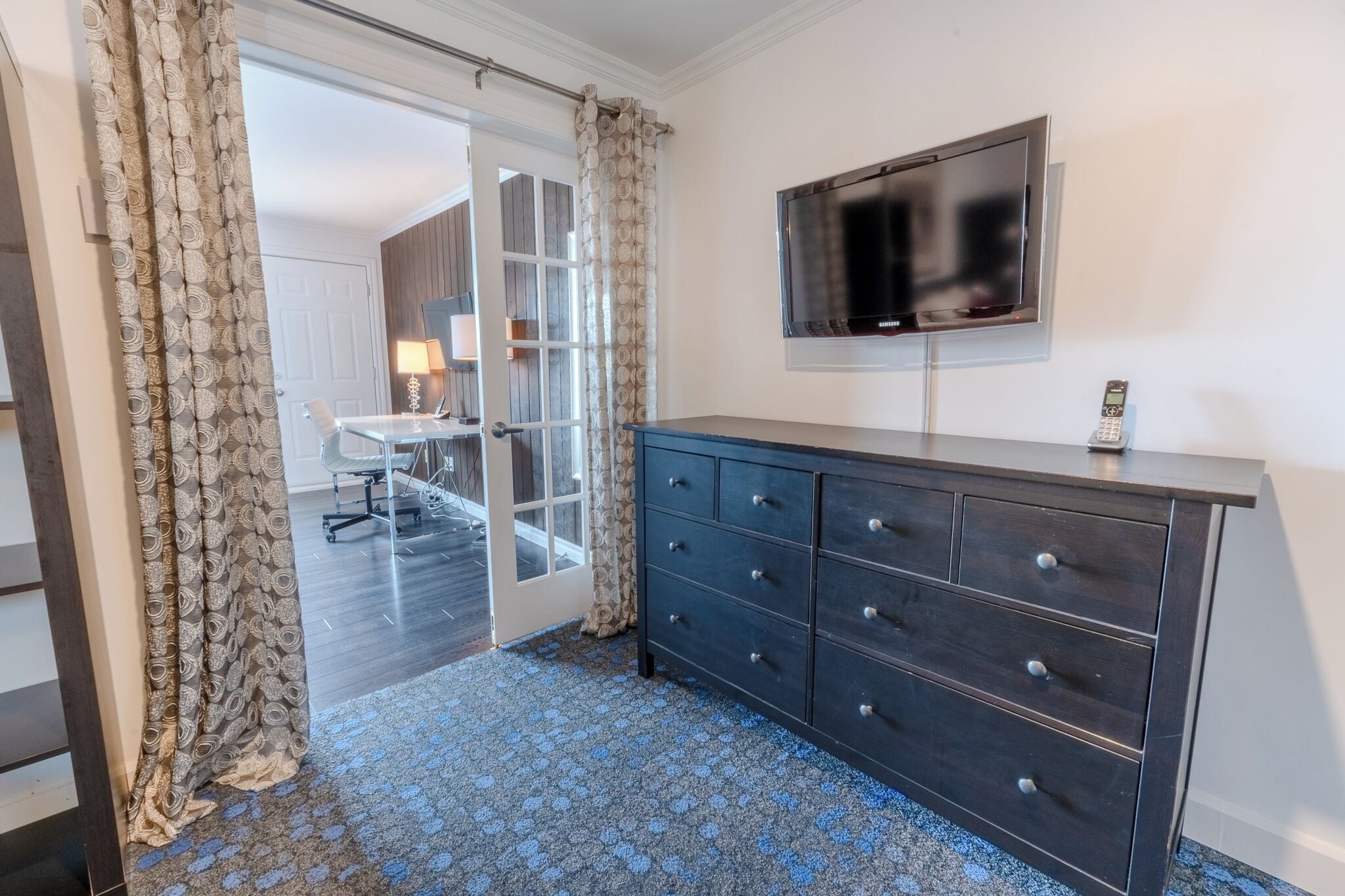 A hotel room with a dark wooden dresser, a flat-screen TV mounted on the wall, and a patterned curtain.