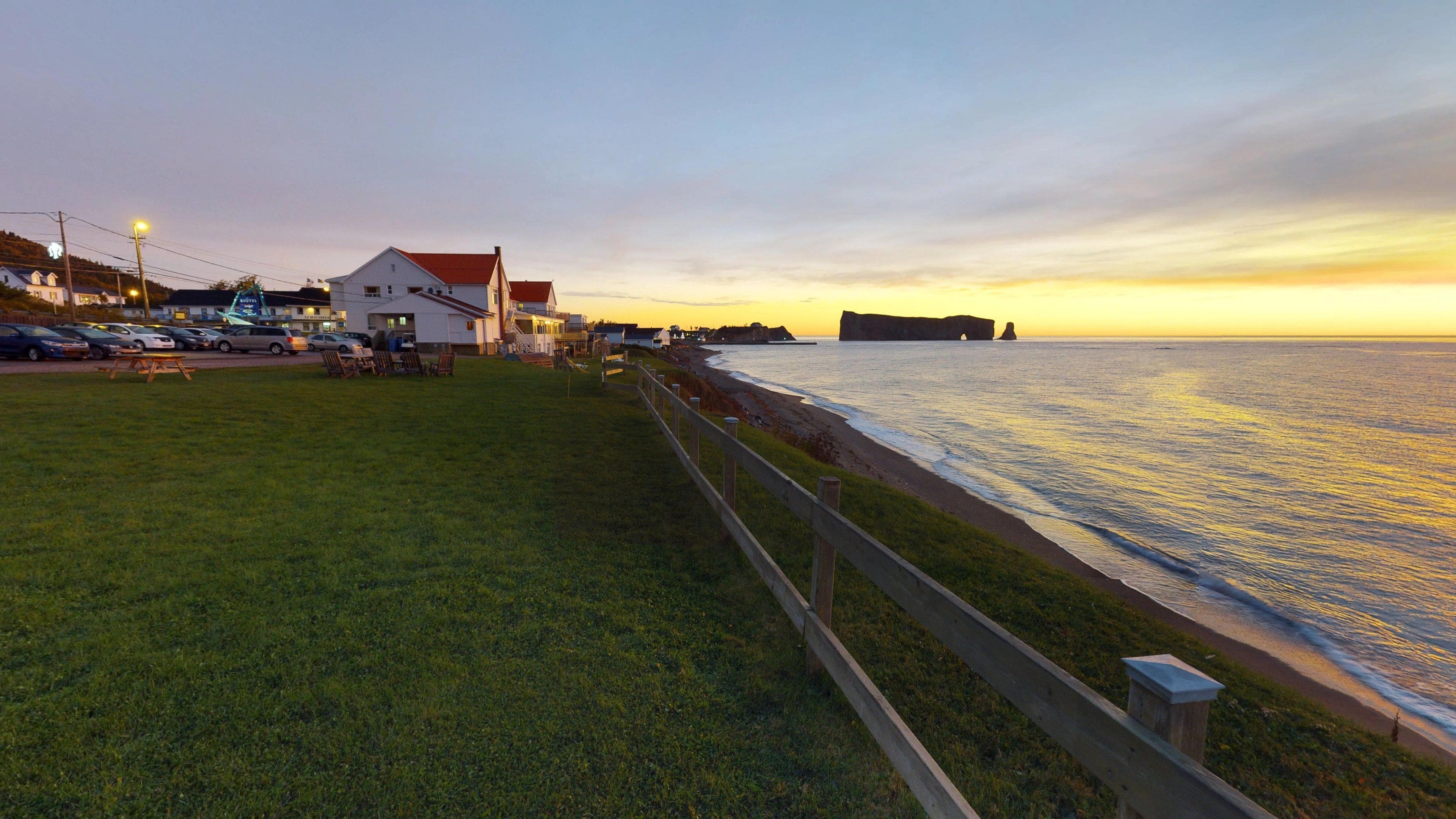 A coastal area with a grassy promenade, a wooden fence, and a building with a red roof.
