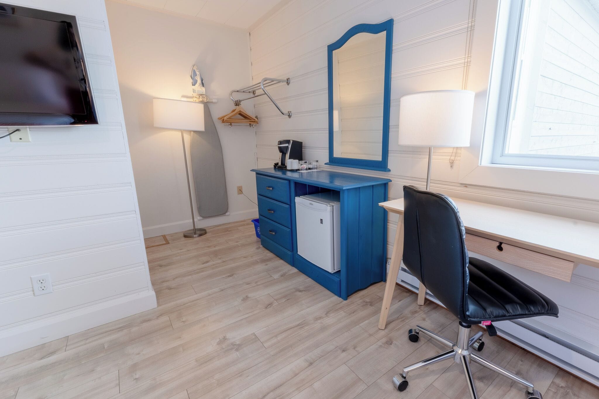 Ein modernes Schlafzimmer mit einer blauen Kommode, einem Schreibtisch, einem Stuhl, einem kleinen Kühlschrank, einem Fernseher und einem großen Spiegel.