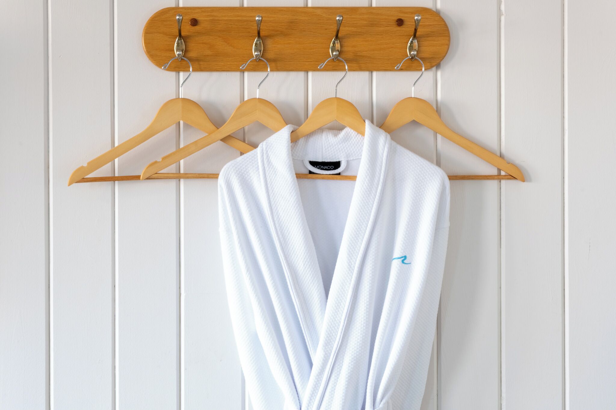 A white robe hanging on a wooden hanger against a white paneled wall.