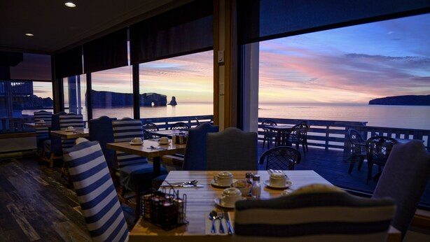 Un restaurante con mesas dispuestas para comer, amplias ventanas que ofrecen vista al agua y la costa distante, y una zona de terraza de madera con sillas.