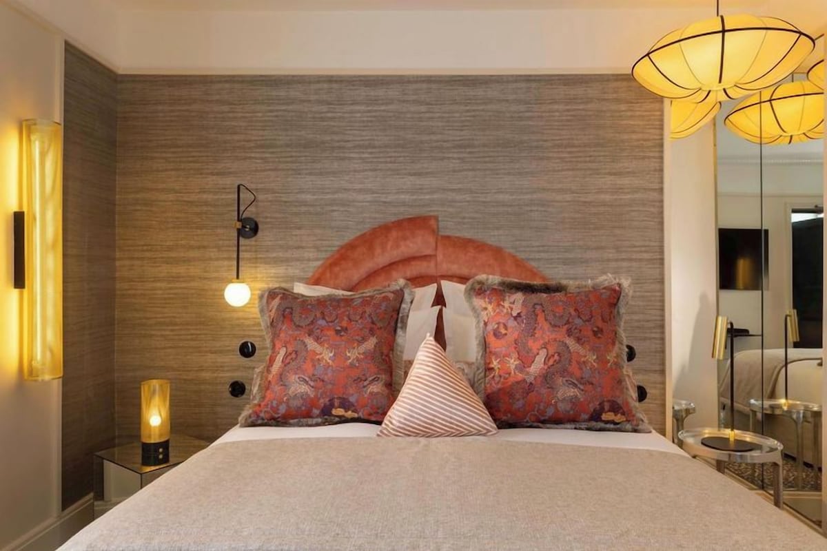 Een moderne slaapkamer met een groot bed, decoratieve kussens, een wandlamp en een nachtkastje met een lamp.