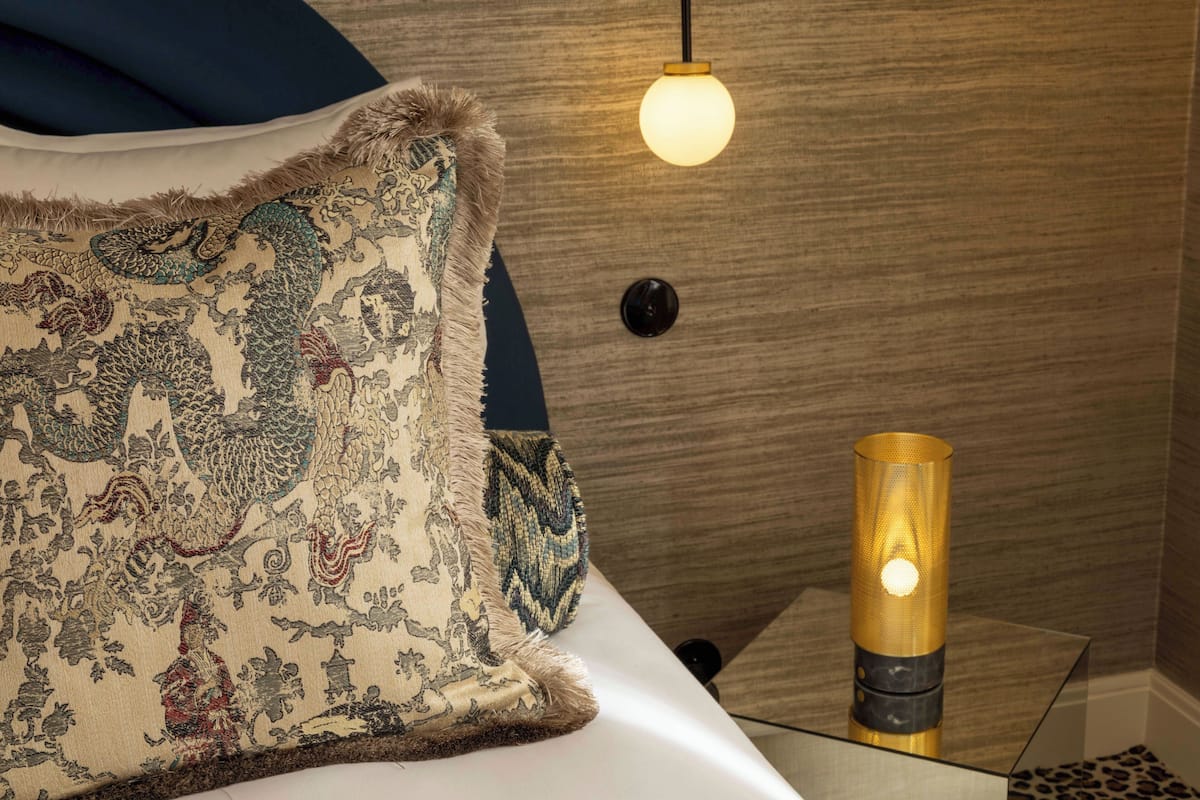 A bed with a patterned pillow, a round wall-mounted light, and a cylindrical lamp on a bedside table.