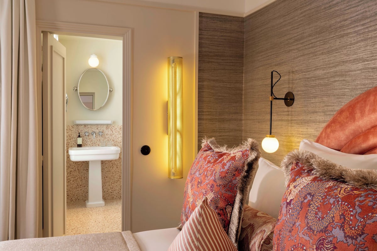 A hotel room with a bed, a bedside table, a lamp, a wall-mounted mirror, and a bathroom visible through an open door.