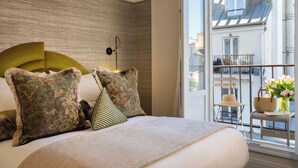 Une chambre à coucher avec un grand lit, une tête de lit et une vue sur un balcon agrémenté d’une chaise en osier et d’un panier.