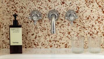Un baño con suelo de terrazo, una botella de gel para cabello y cuerpo Typology y dos vasos vacíos.