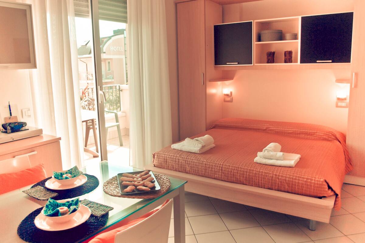 A hotel room with a bed, a desk with breakfast items, a TV, and a balcony with a view of the hotel.