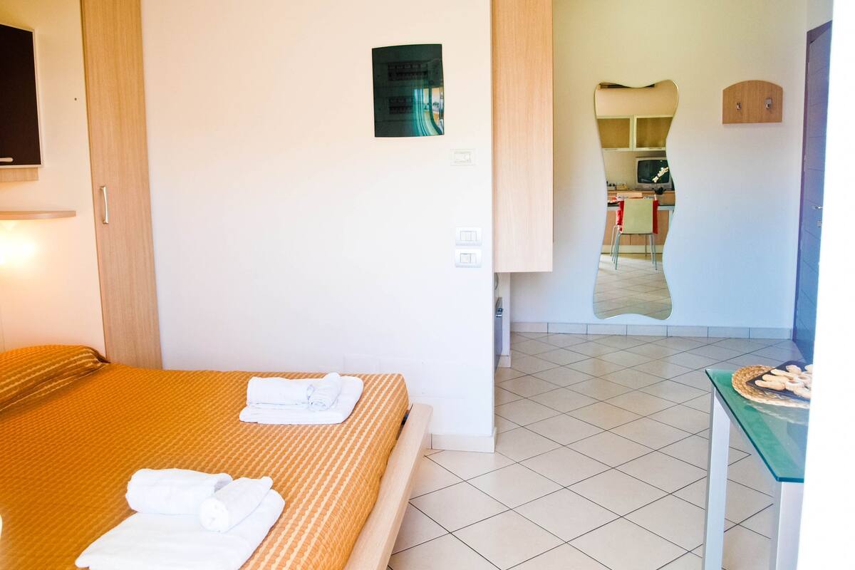 A hotel room with a bed, a small table with a basket, and a kitchenette visible through an open door.