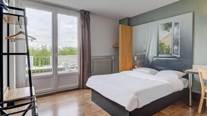 Una habitación de hotel moderna con una cama grande, una silla de madera, una mesita pequeña y vistas al exterior a través de amplios ventanales.