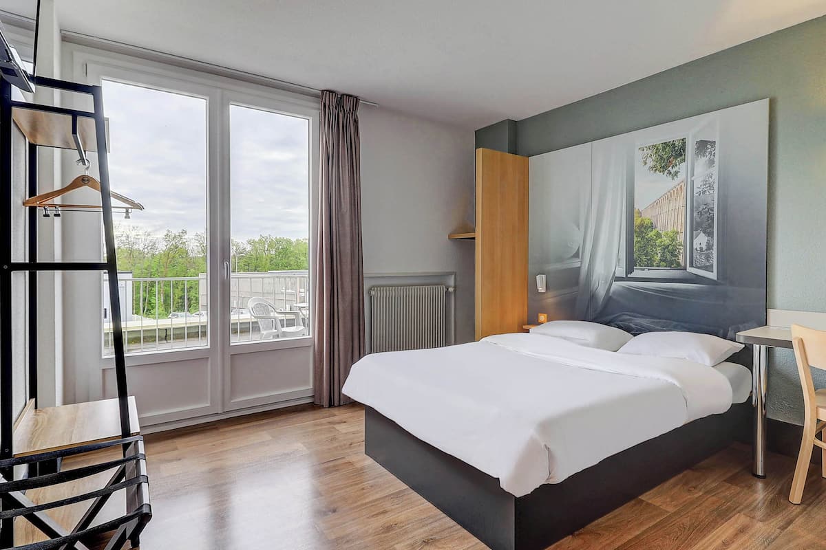 A modern hotel room with a large bed, a wooden chair, a small table, and a view of the outdoors through large windows.
