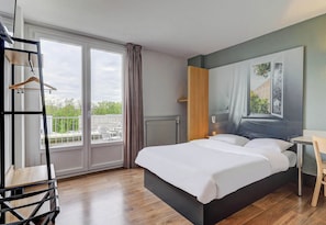 Une chambre d’hôtel avec un grand lit, une chaise en bois, une petite table, un radiateur et une vue sur l’extérieur par une fenêtre.