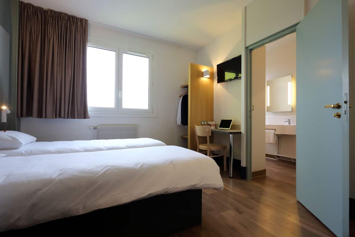 A hotel room with two beds, a desk with a chair, a computer, and a bathroom visible through an open door.