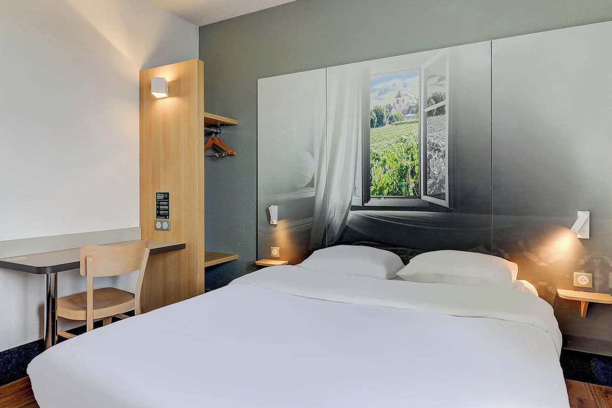 A hotel room with a bed, a desk, a chair, a wardrobe, and a large window with a vineyard view.
