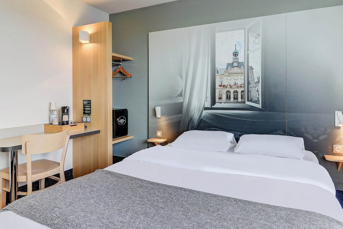 A hotel room with a bed, a desk, a chair, a TV, and a large window with a view of a building.