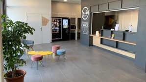 Zona de recepción de hotel con una máquina expendedora, mostrador de recepción y un área de descanso con planta en maceta.