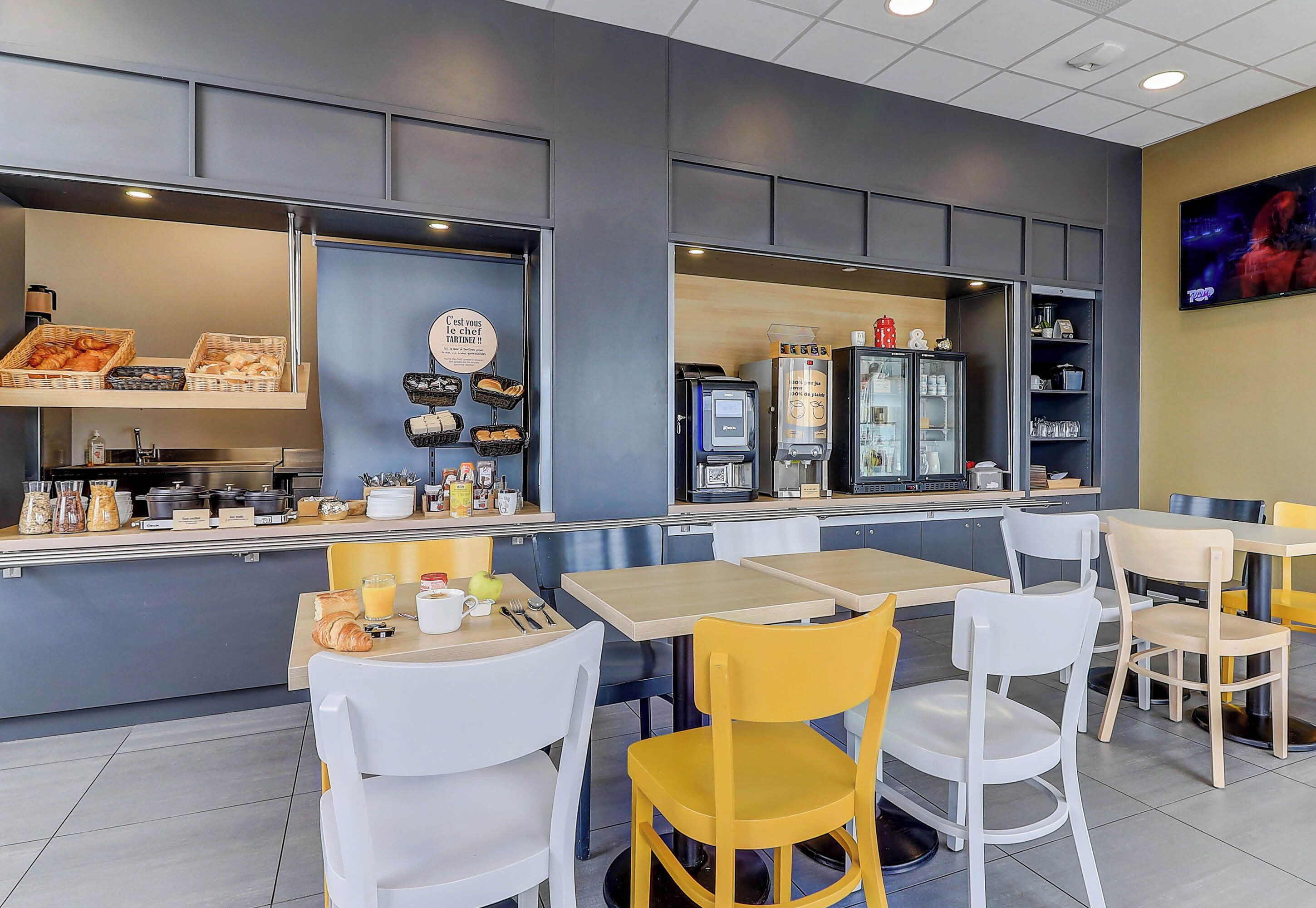 A modern cafe with a counter displaying pastries and coffee machines. There are tables and chairs for seating.