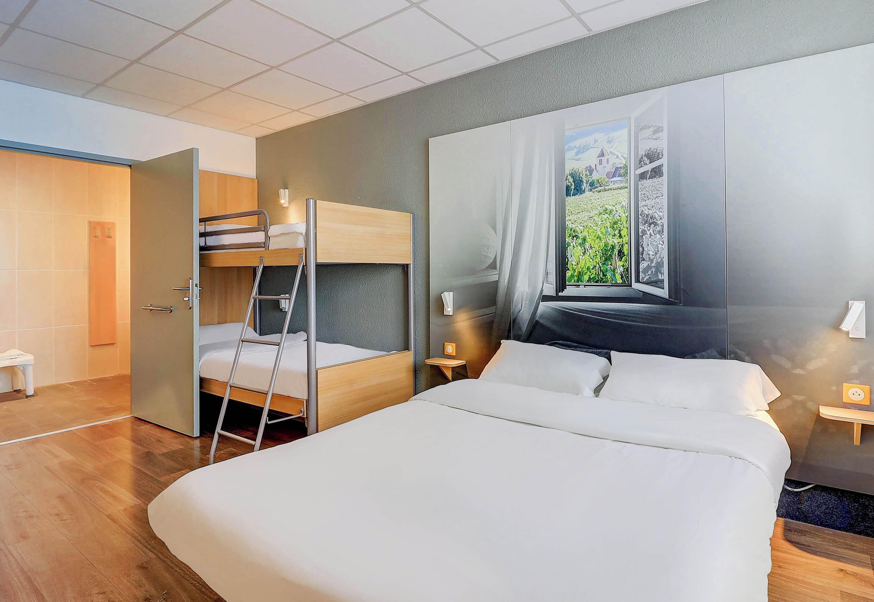 A hotel room with a large bed, a bunk bed, a ladder, a window with a view of greenery, and a small table with a lamp.