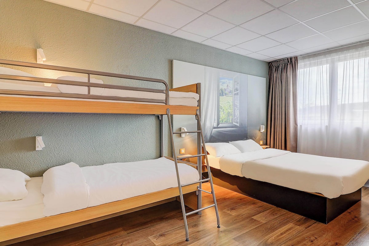 A hotel room with two beds, a ladder, a large window with curtains, and a painting on the wall.
