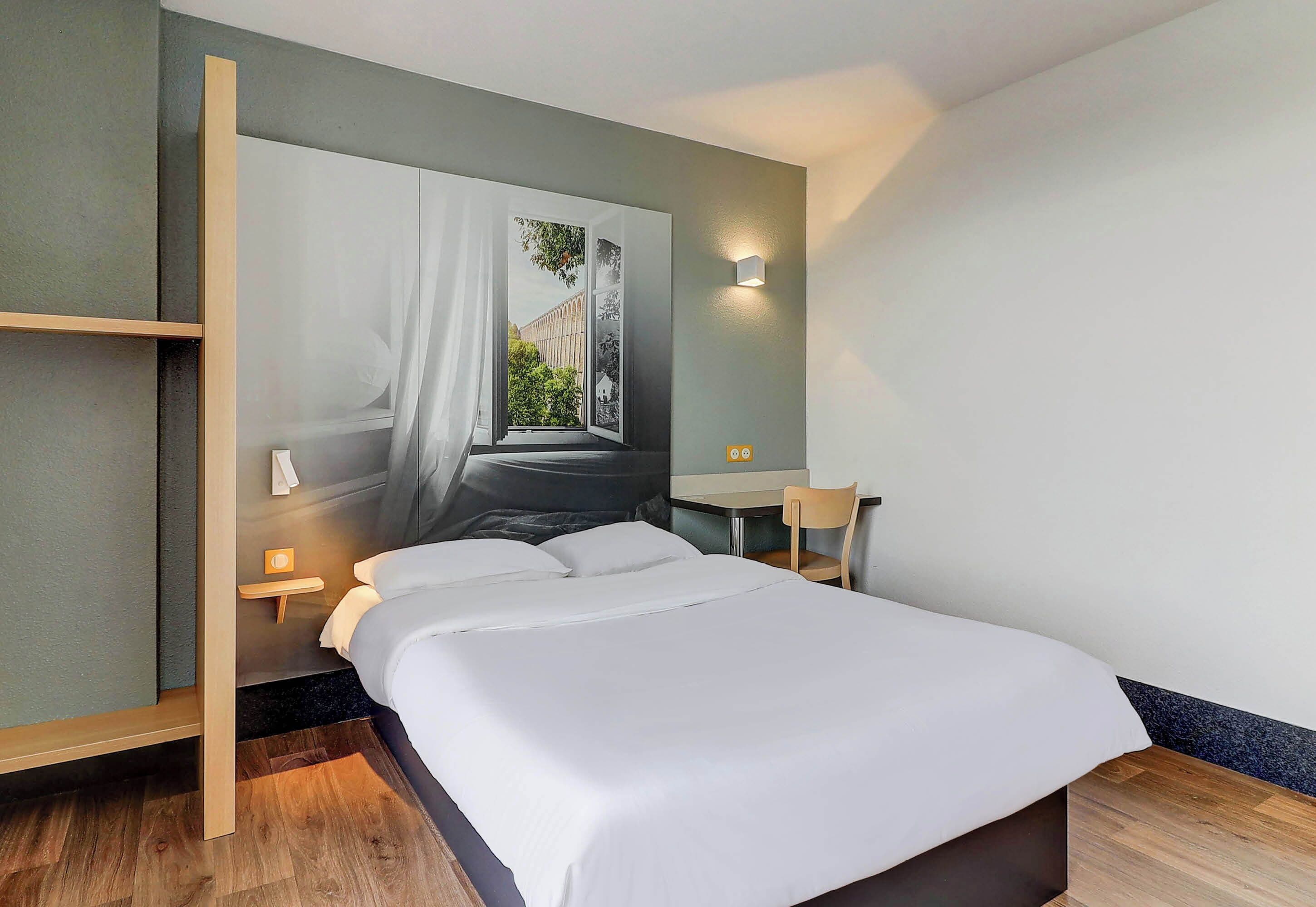 Una habitación de hotel moderna con una cama grande, un escritorio, una silla y vistas a un edificio a través de una ventana.