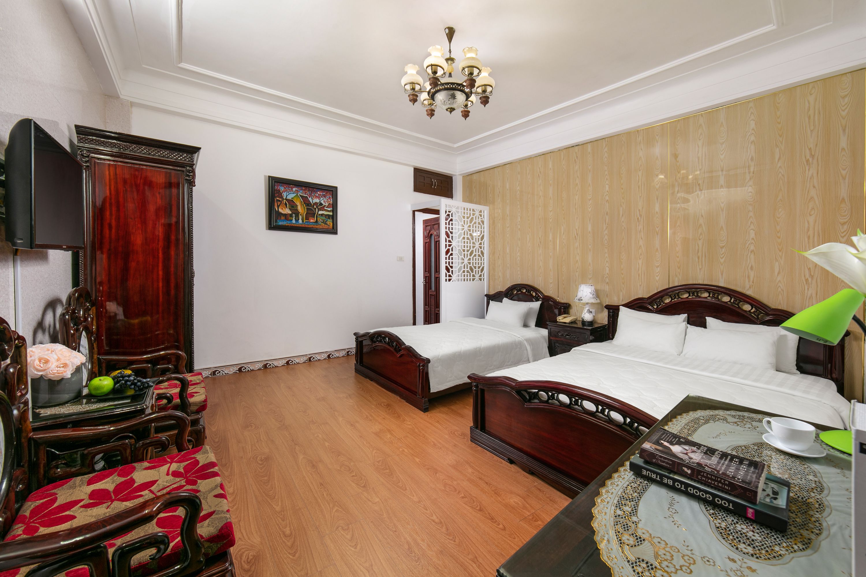 A hotel room with two single beds, a wooden wardrobe, a television, a desk with a lamp, and a decorative screen.