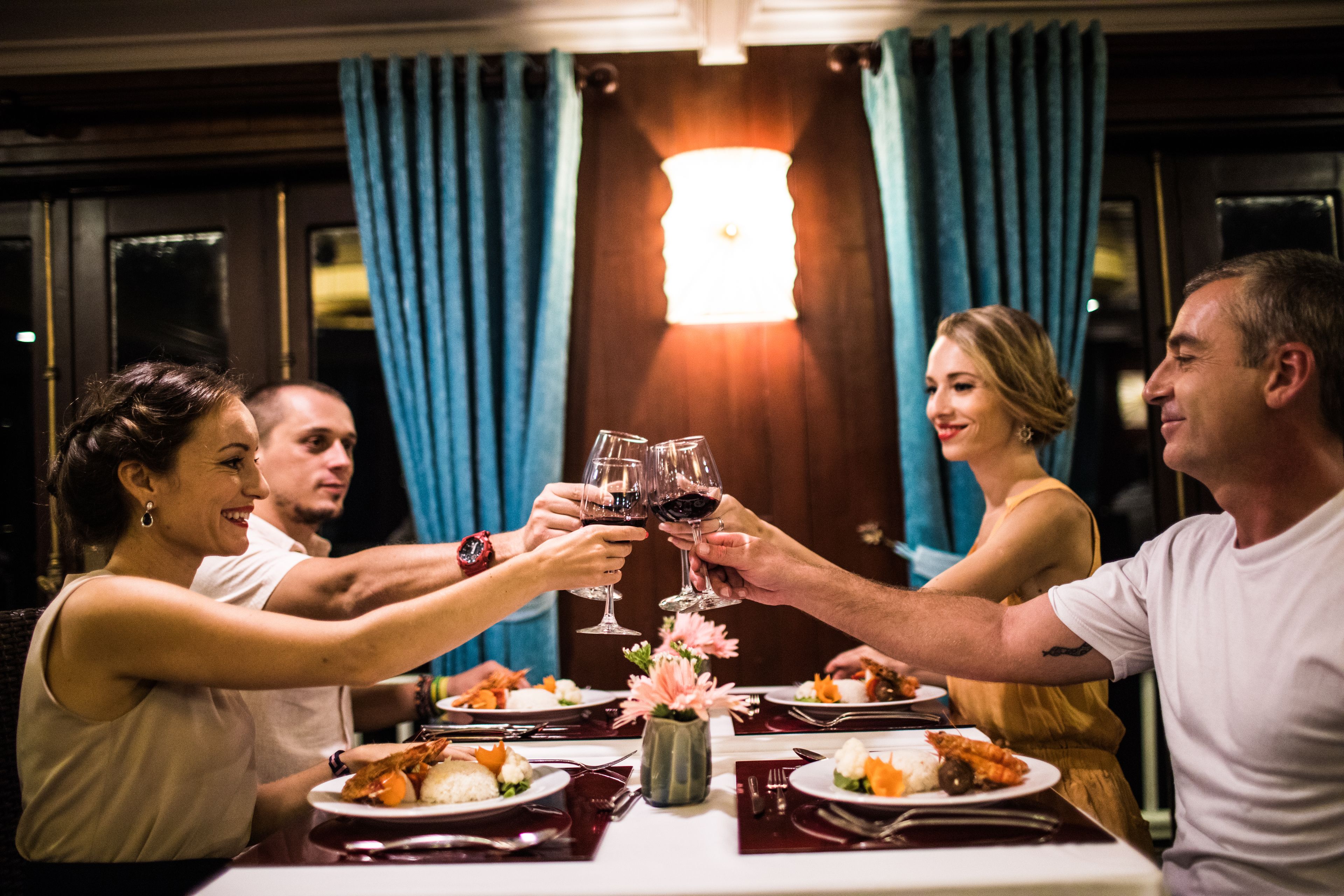 A group of people toasting with wine glasses at a dinner table with plates of food, a vase with flowers, and a lamp in the background.