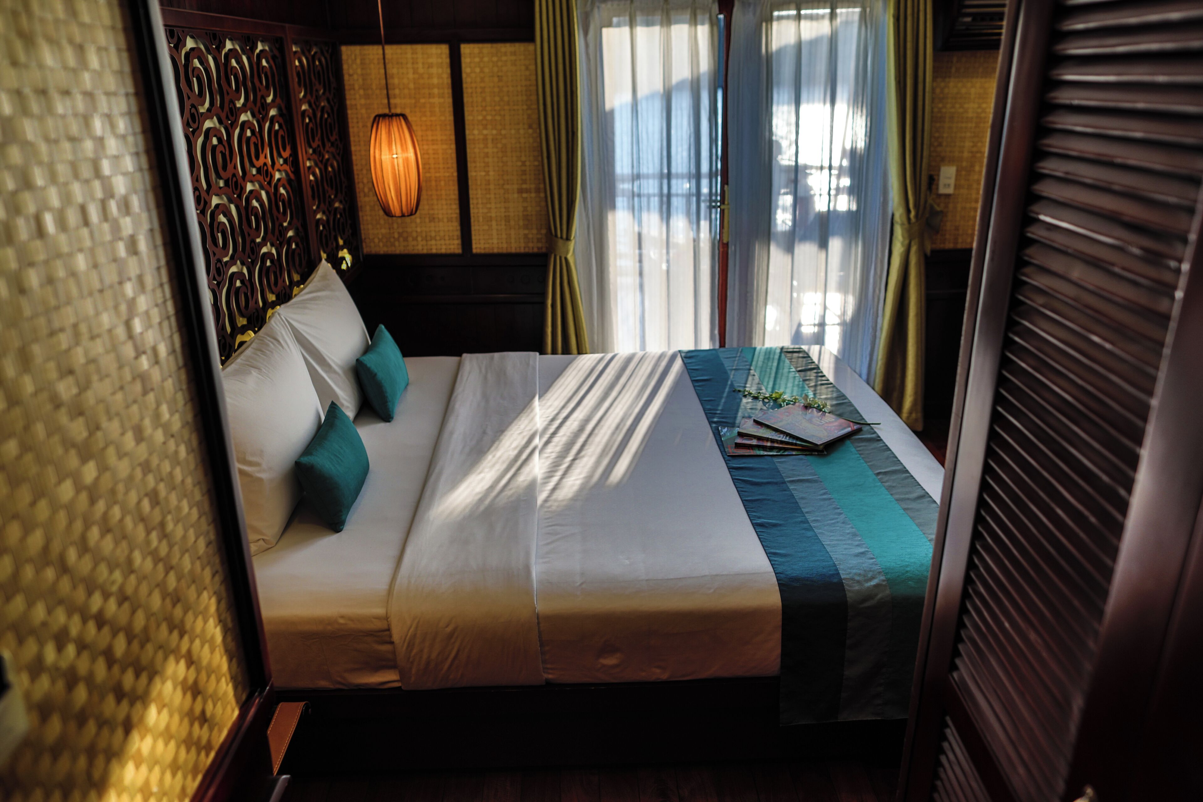 A neatly made bed with white linens and teal pillows, a wooden headboard, and a window with curtains.