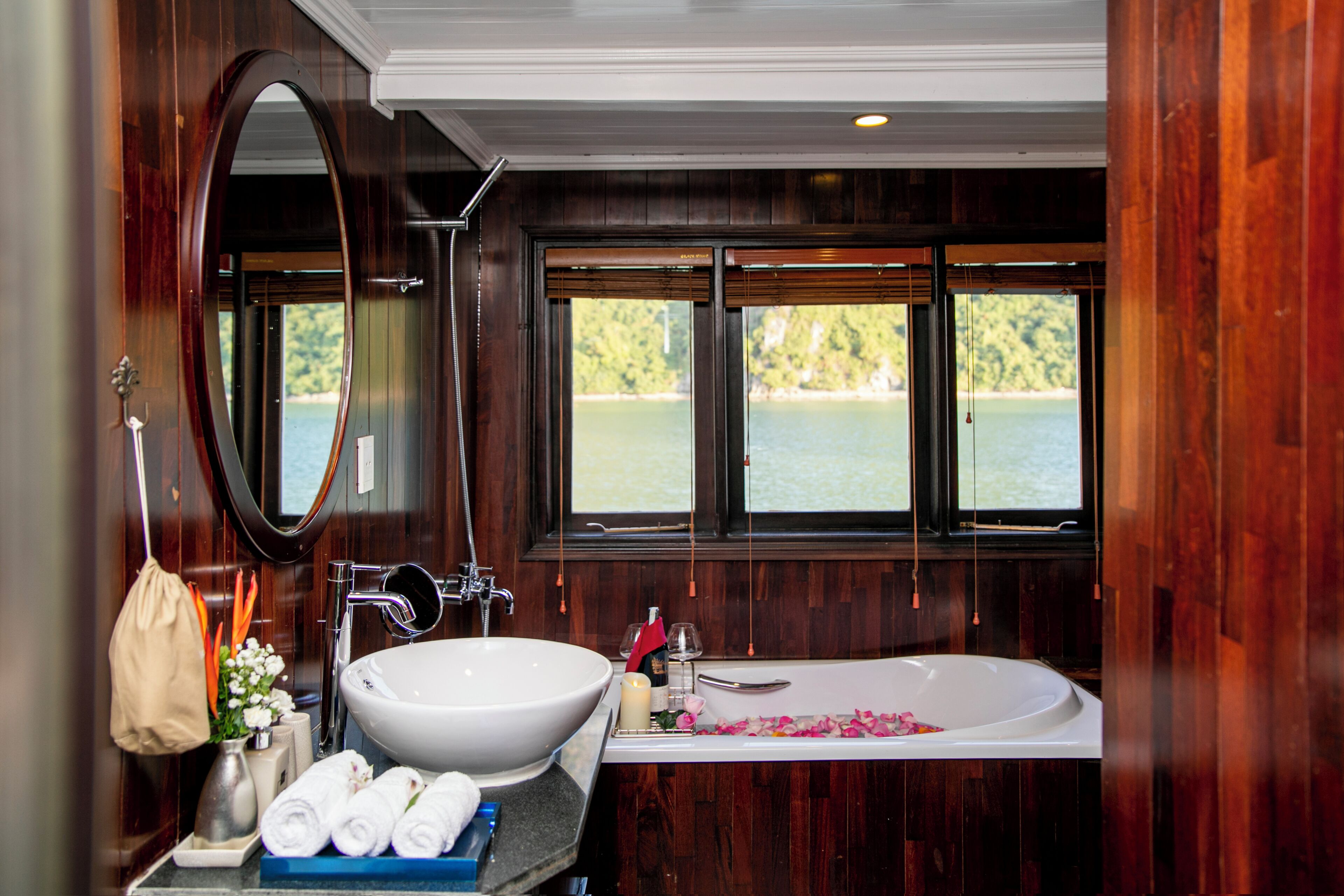 A bathroom with a large oval mirror, a white sink, a bathtub, and a window offering a view of a body of water.