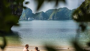 A beach with two people sitting on mats, a boat in the background, and a mountainous landscape.