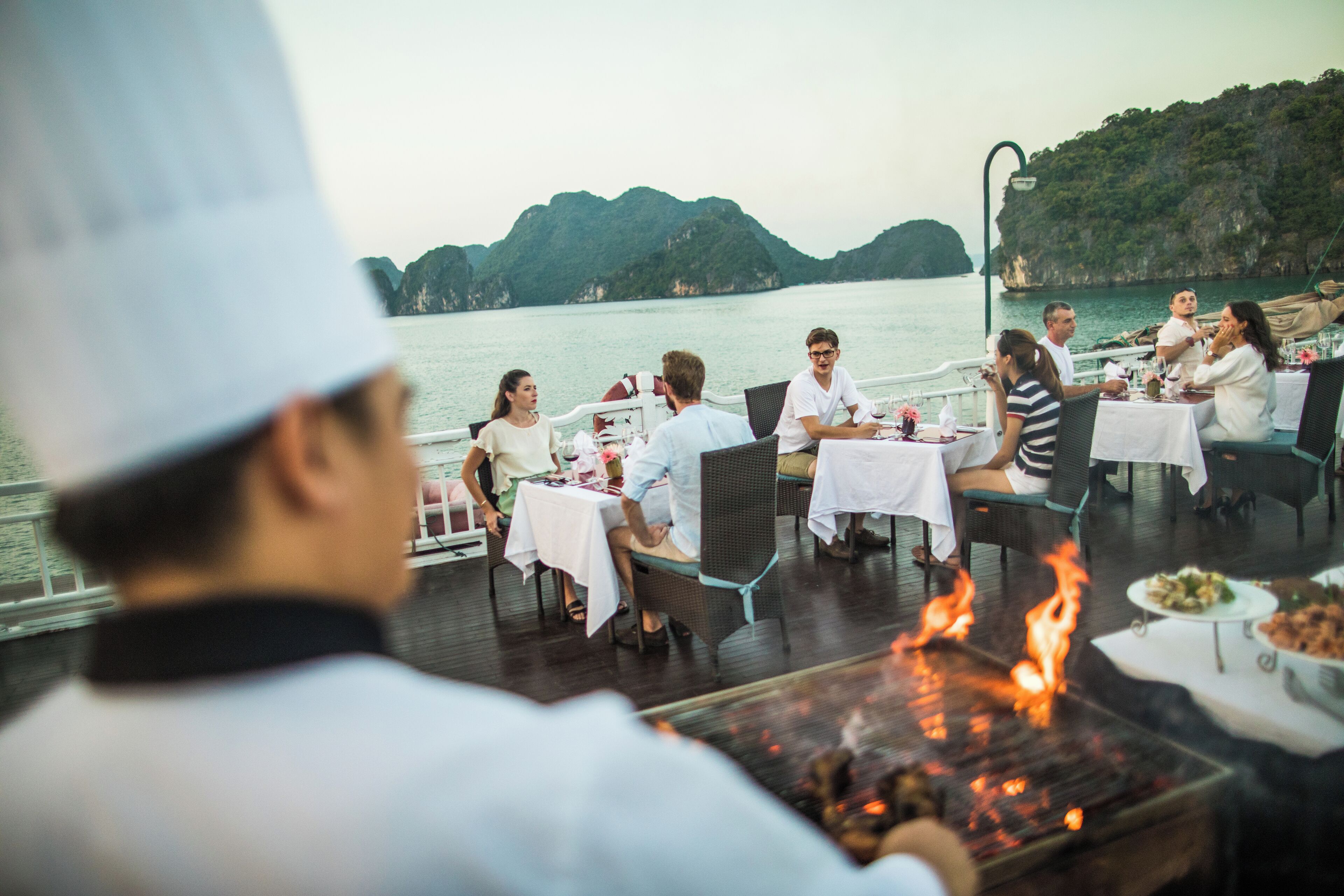 A group of people dining at an outdoor restaurant with a view of the sea and hills.