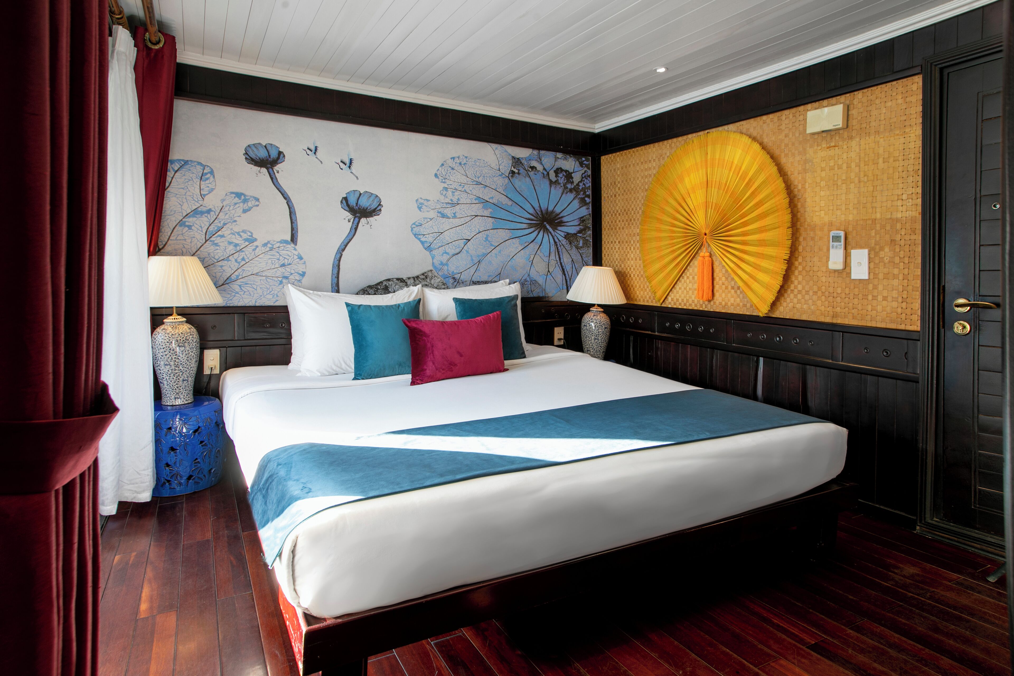 A large bed with a white and blue bedspread, multiple pillows, two bedside lamps, a wall mural, and a large decorative fan.