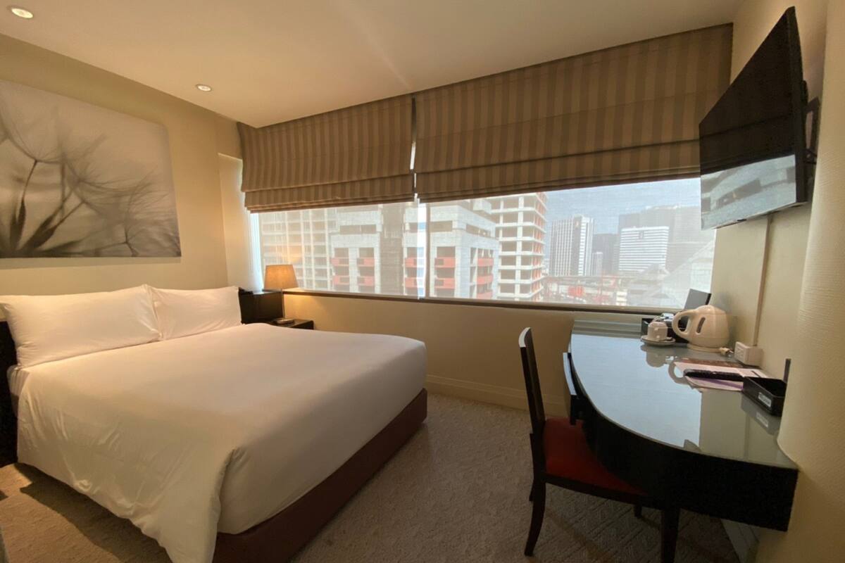 Habitación de hotel con una cama grande, un escritorio con silla, una ventana con persianas y vistas a la ciudad.
