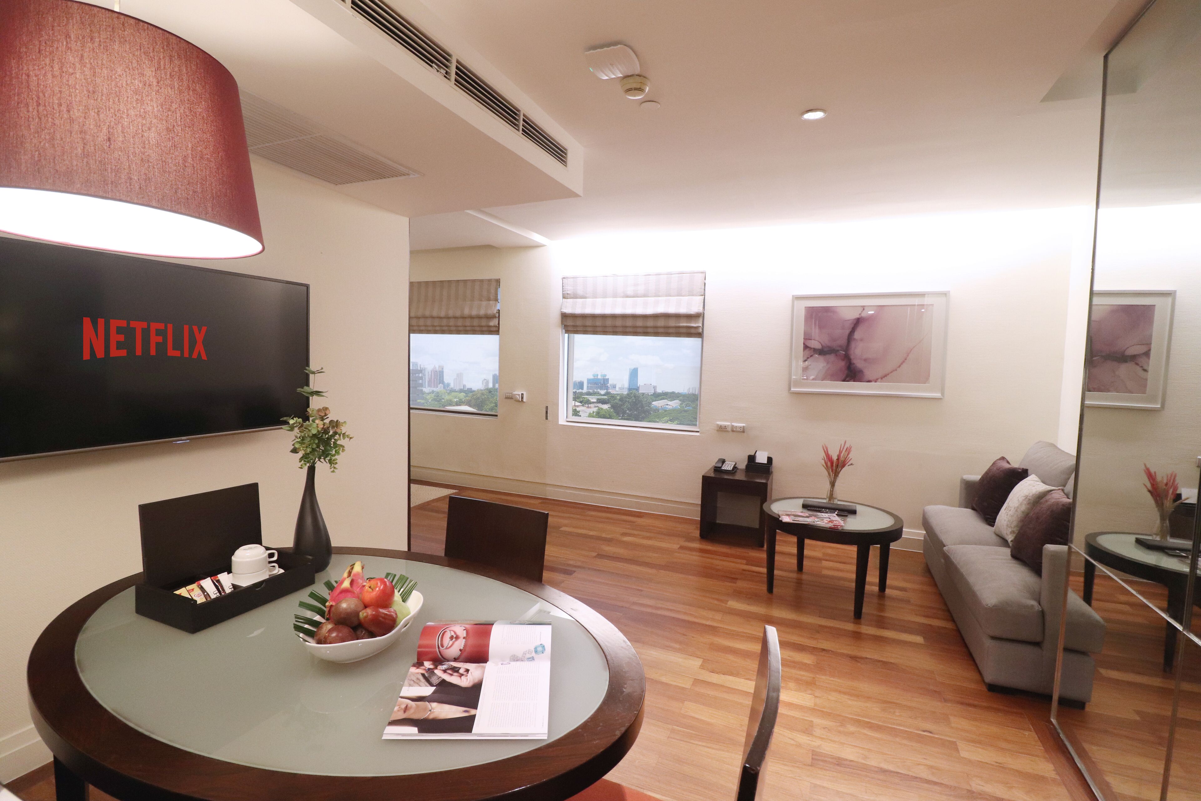 A modern living room with a flat-screen TV, a dining table with a vase and fruit bowl, a sofa, and a city view through large windows.