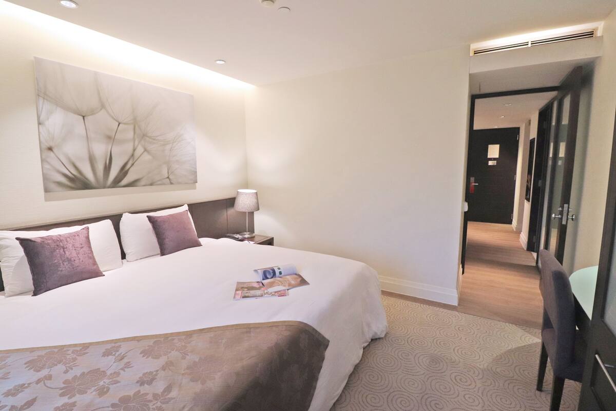 Una habitación de hotel con una cama grande, una mesita de noche con lámpara, una obra de arte en la pared y vistas al pasillo.