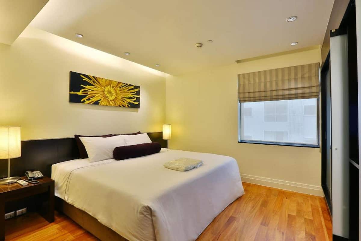 Una habitación de hotel con una cama grande, una mesita de noche con lámpara, un cuadro en la pared y una ventana con persianas.