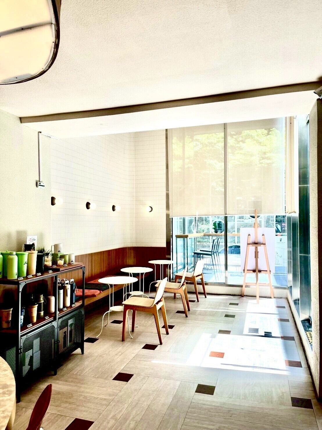A modern cafe with a wooden bench, tables, and chairs. There are large windows providing a view of the outdoors.