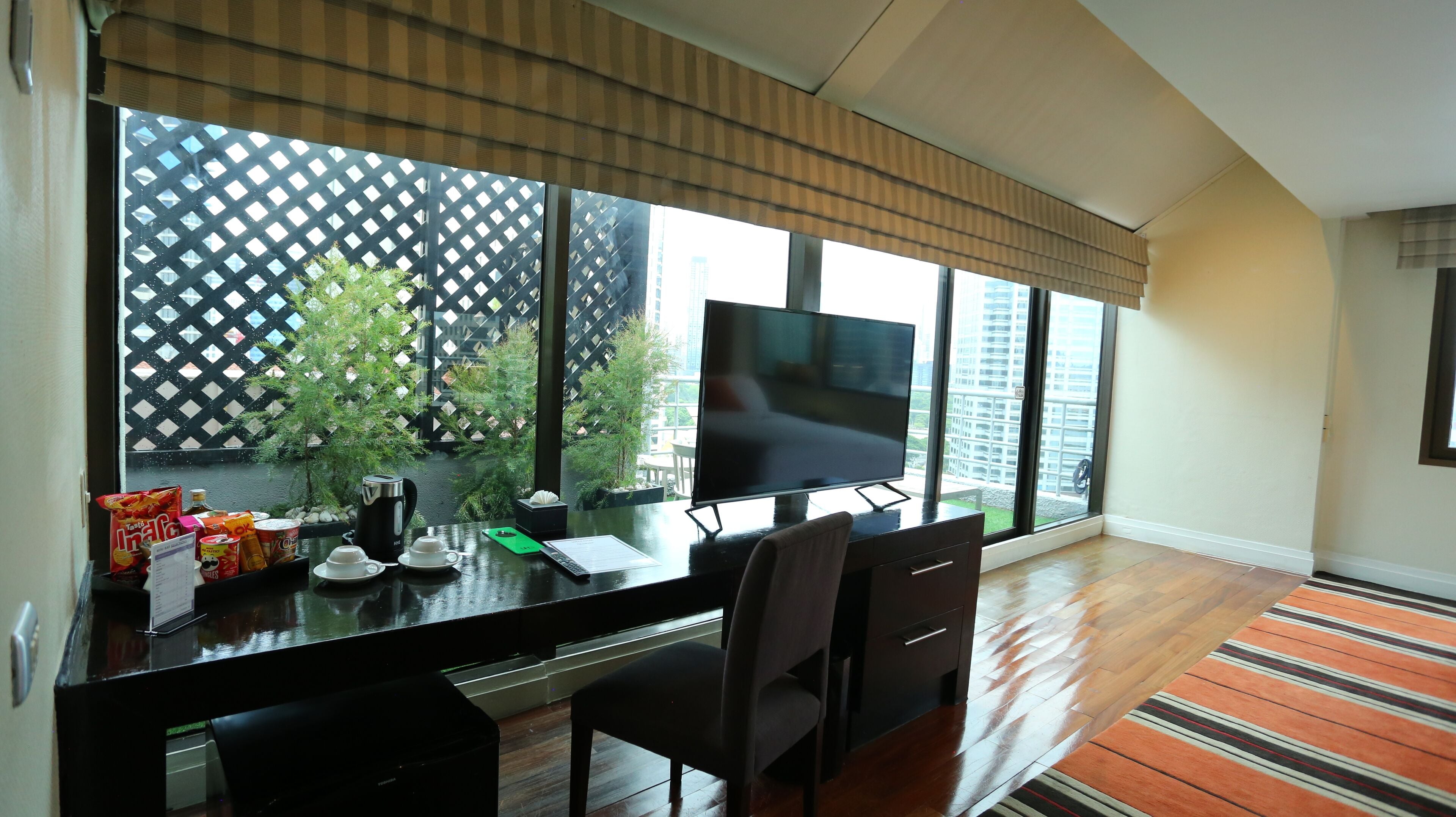 A modern living room with a large TV, a dining table with chairs, a coffee maker, and a view of a cityscape through large windows.